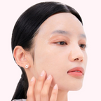 Close-up of a woman with clean, dewy skin wearing a small silver hoop earring, with her hand touching her cheek against a soft light pink background.