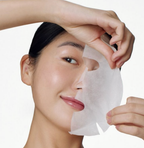 A close-up of a woman applying a translucent white sheet mask to her face against a clean white background.
