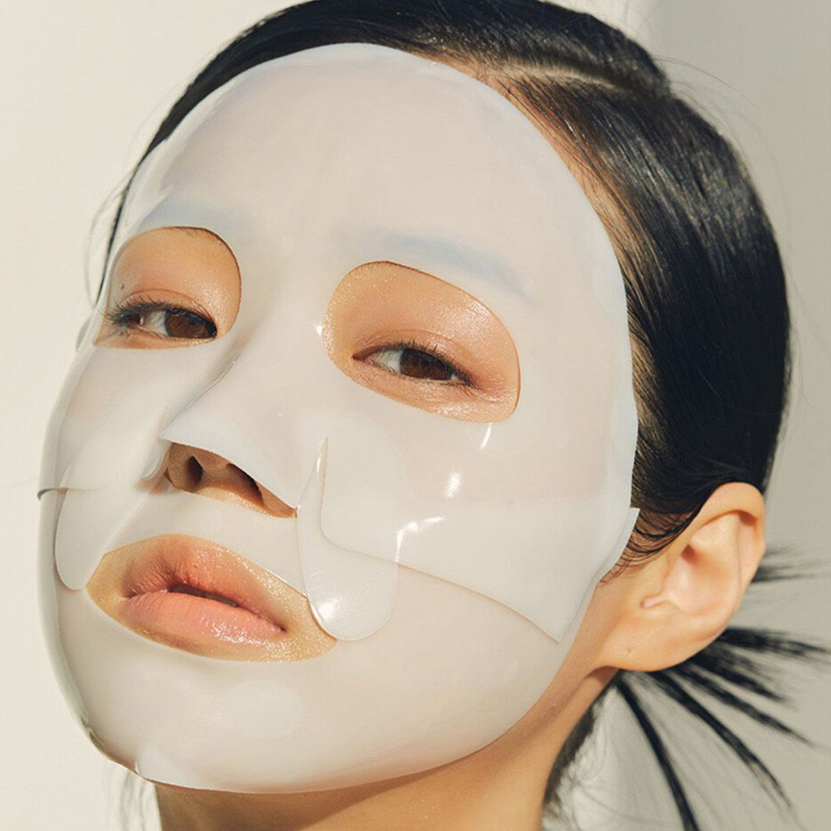 A close-up of a woman wearing a glossy white sheet mask on her face, with her dark hair pulled back, against a plain light background.