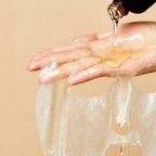 A close-up of a hand holding a translucent, skin-toned sheet mask while a dark bottle pours a golden liquid onto the surface. The background is a solid, warm beige tone.