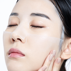 Close-up of a woman with a translucent adhesive eye patch applied to her left eye, touching her cheek with her right hand against a clean white background.