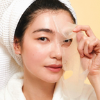 A close-up of a woman wearing a white towel wrap and robe, peeling a translucent beige sheet mask from her cheek against a soft yellow background.