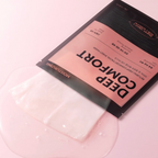 A sheet mask packet labeled 'DEEP COMFORT' with a translucent white mask partially peeled back, set on a glossy pink surface.