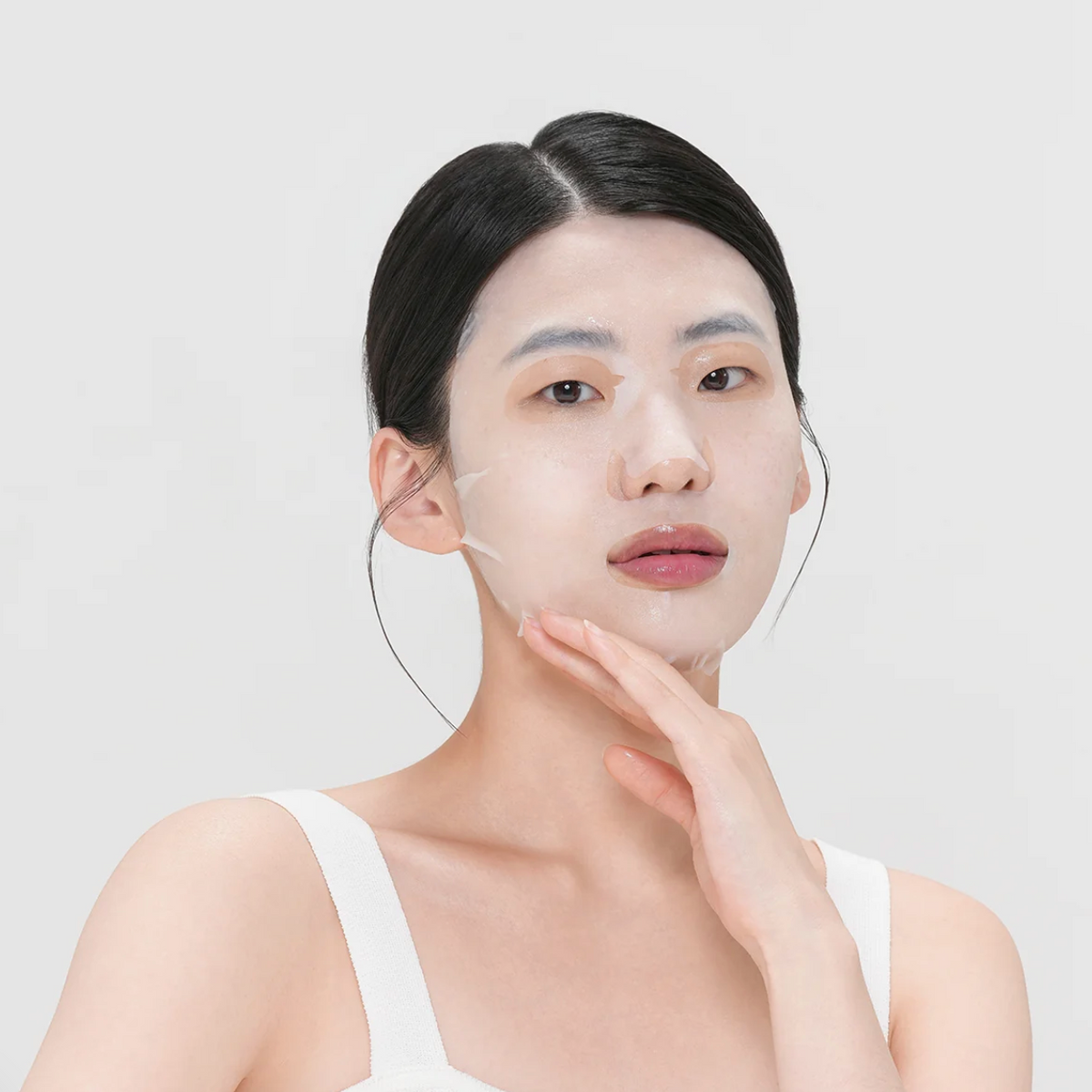A woman with dark hair pulled back wears a translucent peel mask on her face, touching her cheek against a clean, light gray background.