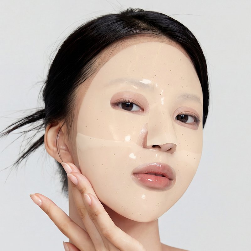A close-up of a woman with a glossy, translucent beige facial mask or liquid makeup applied to her face, touching her cheek with her hand against a clean, light gray background.