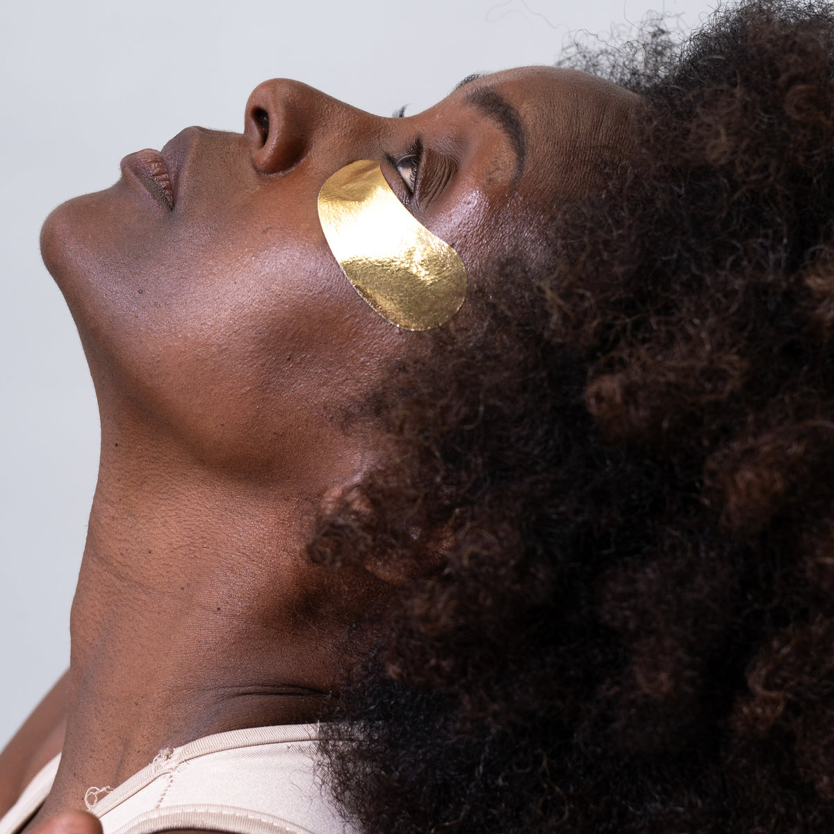 A close-up of a woman with dark curly hair wearing a shiny metallic gold under-eye patch against a light gray background.