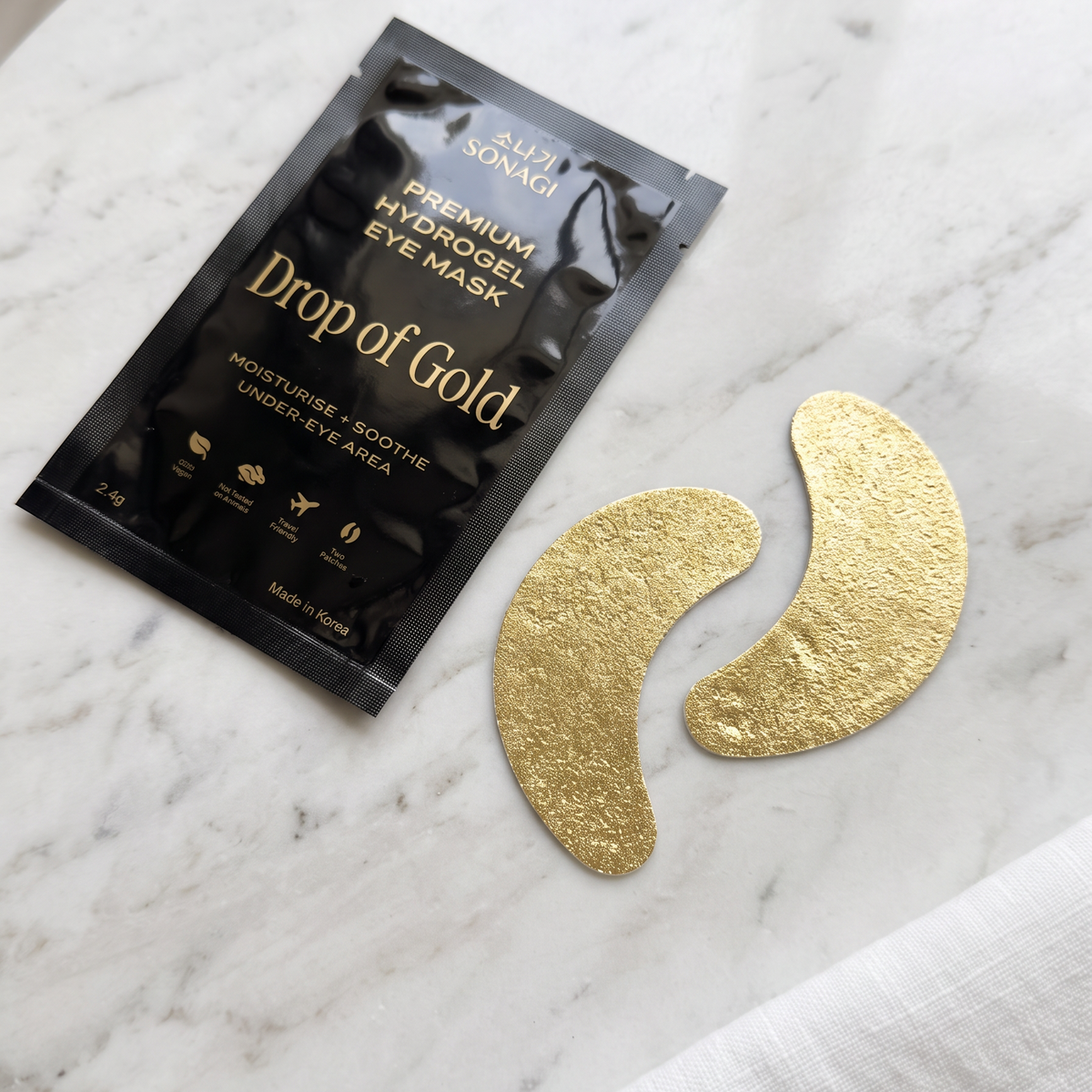 A black packet of Gold Nomad 'Drop of Gold' premium hydrogel eye mask lies on a white marble surface next to two gold crescent-shaped patch masks and a folded white cloth.
