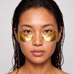 A close-up of a woman with wet dark hair wearing two gold eye mask patches under her eyes, set against a plain light gray background.